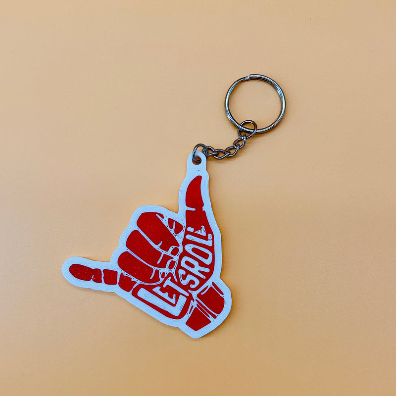 A red and white 3d printed keychain shaped like a hand gesture with the word 'Lets roll' in white, attached to a silver metal split ring, set against a solid peach background.