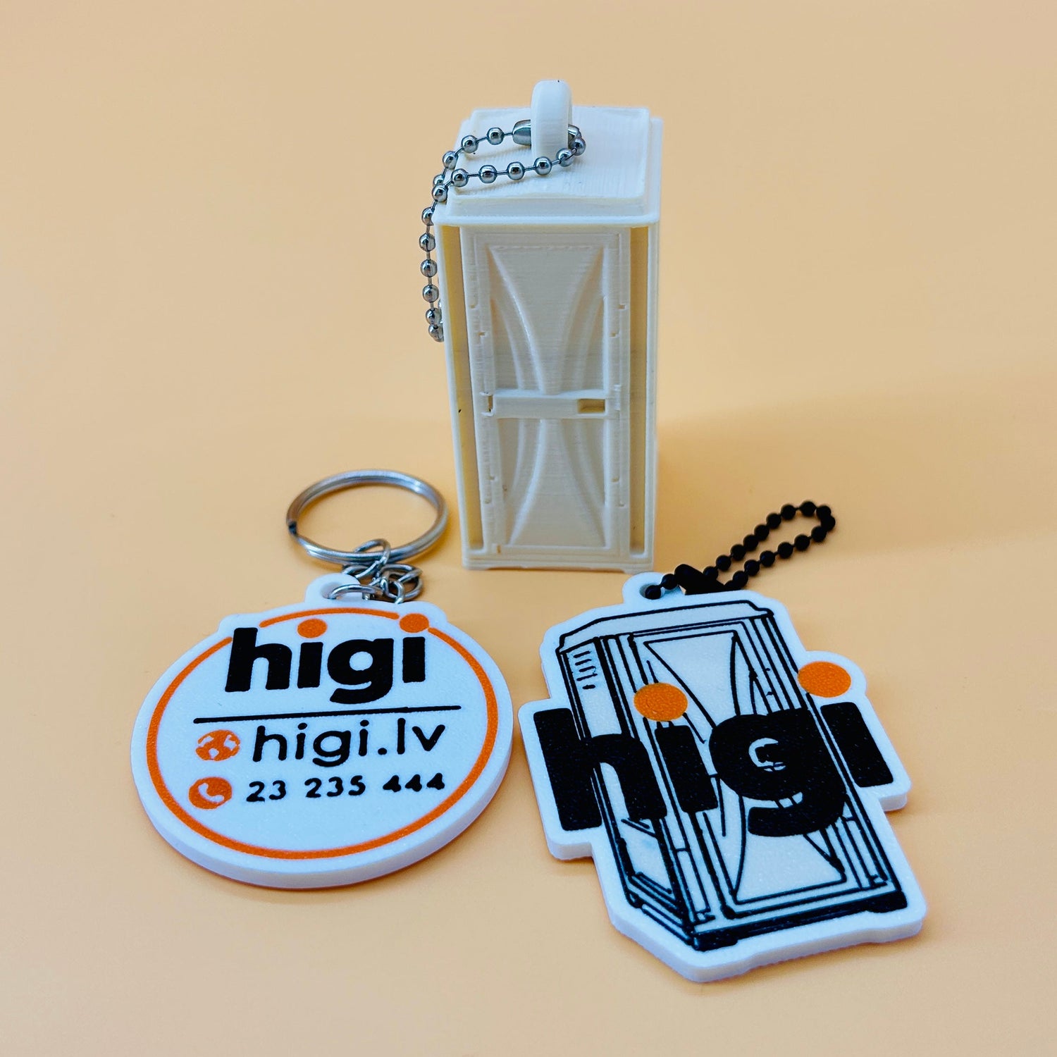 Three custom 3d printed keychains with a white bio toilet booth charm and printed designs for 'higi' and 'higi.lv' on a warm beige background.