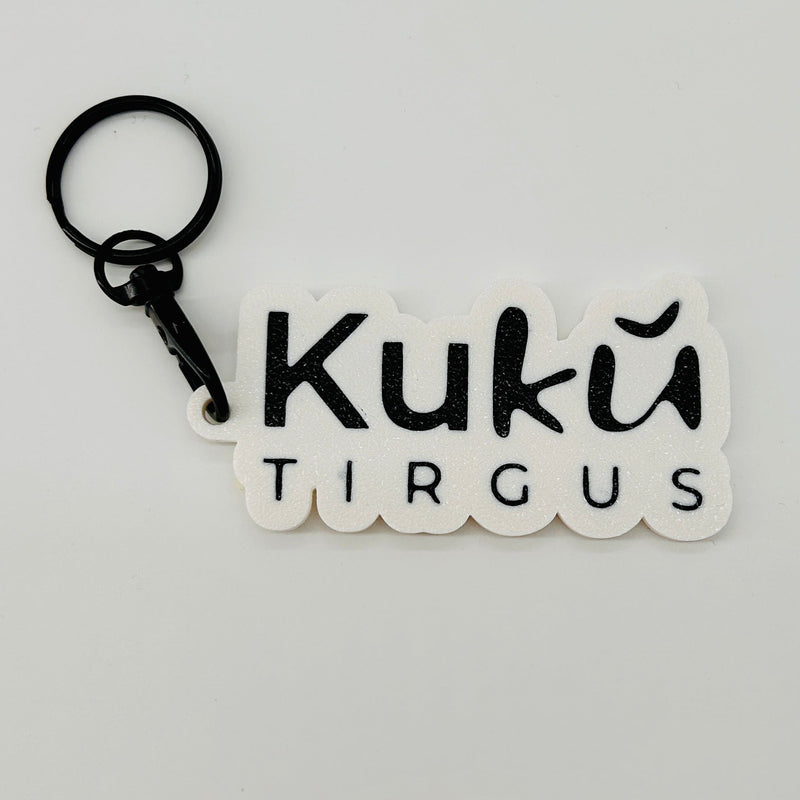 A white 3d printed keychain with the words 'Kukû TIRGUS' in black lettering, attached to a black split ring, on a plain light gray background.